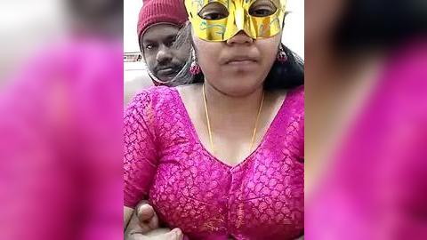 Media: Video of a woman with medium brown skin, wearing a pink lace blouse and a golden Venetian mask, holding a dark-skinned man's hand, blurred in the foreground.