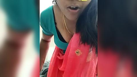 Media: Video of a woman with medium skin tone and long black hair, wearing a teal shirt and a red saree with a gold floral design. She's leaning forward, her mouth open as if speaking.