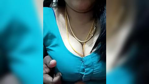 Media: Video of a woman in a bright turquoise top, wearing multiple gold necklaces and a gold chain. Her breasts are prominent. Blurry background, likely an indoor setting.