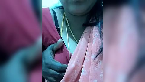 Media: Video showing a close-up of a woman with medium skin tone and long black hair, wearing a pink sari and a white blouse, with a gold necklace, hands positioned under the blouse, blurred background.