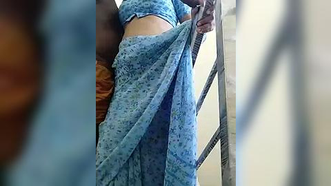 Media: Video of a person wearing a blue, floral-patterned saree, climbing a metal ladder in a cluttered room with a brown sofa and beige walls.