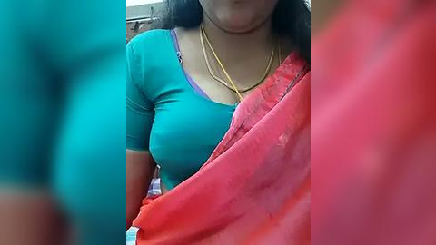 Media: Video of a South Asian woman with medium skin tone, wearing a teal t-shirt and a red sari with a gold necklace. Background is blurred.