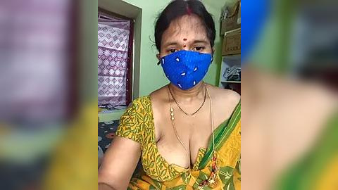 Media: Video of an Indian woman with medium skin tone, dark hair, and a blue face mask, wearing a yellow sari with floral patterns. She has a gold necklace and is indoors with a green wall and a window with a patterned curtain.