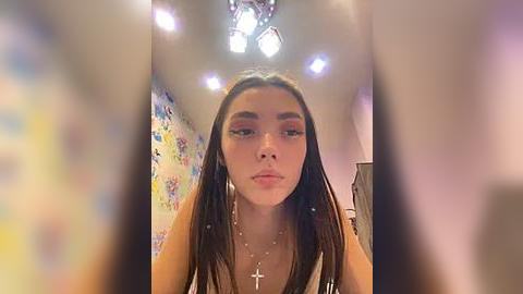 Media: A video of a young woman with long dark hair, wearing pink eyeshadow, a cross necklace, and a sleeveless top, standing in a colorful, whimsical room with pastel walls and a chandelier.