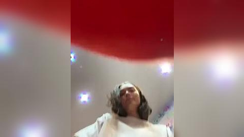 Media: A video of a young woman with shoulder-length hair and a white shirt, captured from a low angle, showing a red and white gradient background with blurry lights.