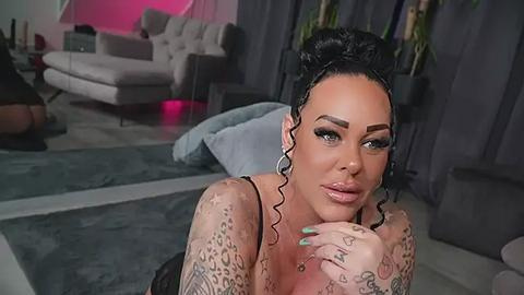 Media: Video of a tattooed woman with dark hair styled in a bun, wearing hoop earrings, and black lingerie, leaning against a gray couch in a modern living room with gray curtains and a beige sectional.