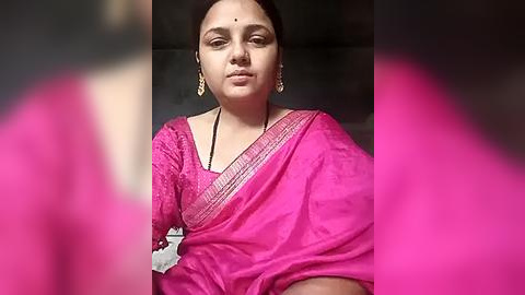 Media: Video of a woman in a bright pink saree with intricate gold embroidery, wearing large, gold earrings, and a white blouse. Her facial expression is calm, and she has a small dot on her forehead. The background is blurred.