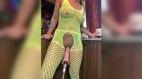 Media: Video of a woman in a neon green fishnet bodysuit, revealing her genitals, holding a pink dildo. Background features a blurred Christmas tree and wooden cabinets.