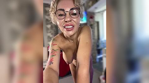 Media: Video of a topless woman with blonde hair and glasses, smiling, showing tattoos on her arm. She's indoors, blurry background.