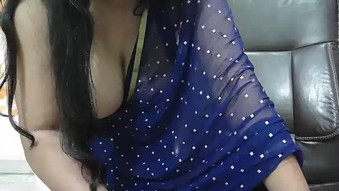 Media: A video of a woman with long black hair, wearing a sheer blue blouse with white polka dots, revealing her large breasts and a green bra underneath, seated on a brown leather chair.