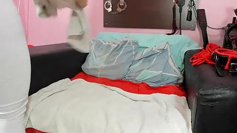 Media: Video of a person in a black leather jacket and white pants holding a white pillow in a pink room with handcuffs on a wall, a bed with a red blanket, and a black couch.