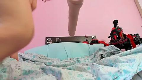 Media: Video of a woman's bare breast, partially covered by a blue and white patterned blanket, in a room with a pink wall, a Harley-Davidson motorcycle, and a large white dildo hanging from the ceiling.