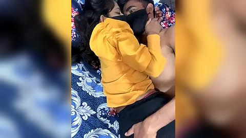 Media: A video shows a woman with long black hair in a yellow top, embracing a shirtless man with medium skin tone on a blue and white floral bedspread.