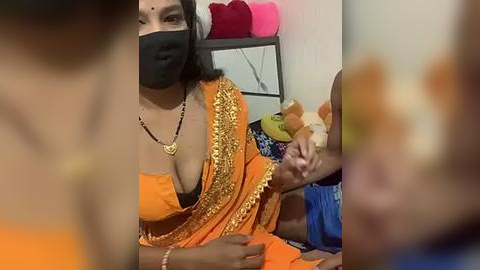 Media: Video of a woman in an orange sari with gold embroidery, wearing a black mask, holding a stuffed toy in a room with a red pillow and a shelf.