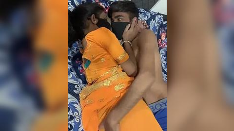 Media: Video of an intimate moment between a topless man and a woman in an orange sari, both wearing face masks, lying on a blue and white patterned bedspread.