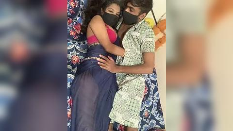 Media: A video shows a couple, both masked, lying on a bed with floral sheets. The woman, in a sheer blue dress, is embracing the man, who wears a patterned shirt. The room is blurred.