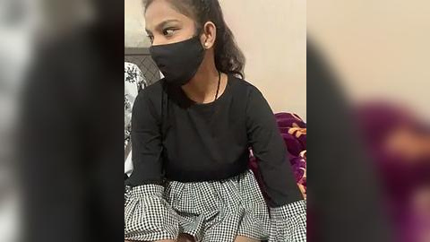 Media: Video of a young South Asian woman with medium skin tone, wearing a black face mask, black long-sleeve shirt, and checkered skirt, sitting in a room with a purple blanket and beige walls.