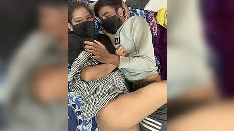 Media: Video of two men, one in a gray hoodie, the other in a black shirt, lying closely together on a bed with patterned sheets. They wear black masks and are in a cozy, intimate setting.