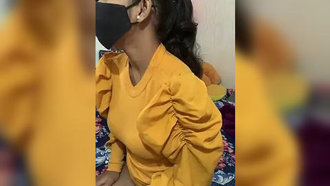 Media: A video of a woman with medium skin tone, wearing a mustard-yellow blouse with ruffled sleeves, a black mask, and glasses, seated on a bed with a colorful, floral-patterned blanket.