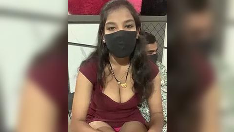 Media: Video of a young Asian woman with long black hair, wearing a black face mask, maroon top revealing cleavage, and pink shorts, sitting indoors with a blurred background.