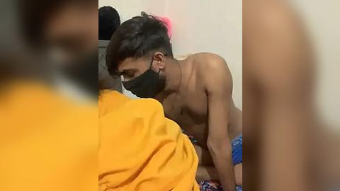 Media: Video of a shirtless young man with dark hair, wearing a black mask, performing oral sex on a person in a yellow shirt, both seated on a bed with blue-patterned sheets.