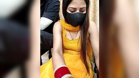 Media: A video of a South Asian woman in a yellow saree, black mask, and red bangles, seated on a chair.