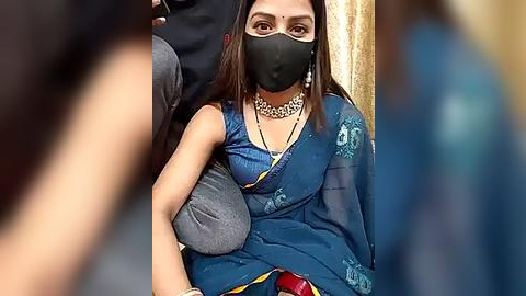 Media: Video of a South Asian woman with medium skin tone, wearing a blue sari, black mask, and gold necklace, seated on a grey cushion, blurred people in the background.