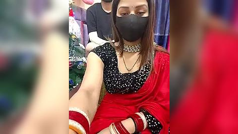 Media: Video of a woman in a red sari with a black blouse, black face mask, and gold choker, seated in a crowded indoor setting.