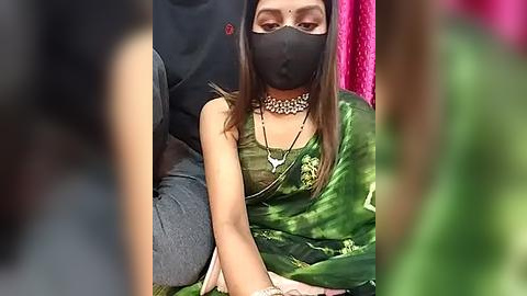 Media: Video of a woman wearing a black mask, green saree with gold embroidery, black choker, seated on a pink background, with blurred figures in the foreground.