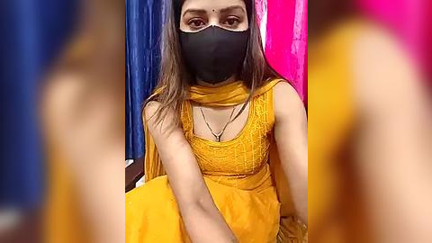 Media: Video of a South Asian woman with long brown hair, wearing a yellow embroidered kurta and a black mask, seated against blue and pink curtains.