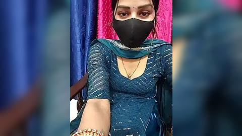 Media: Video of a woman with light brown skin, wearing a black face mask, teal embroidered dupatta, and blue shirt. She sits on a pink and blue cushioned chair.