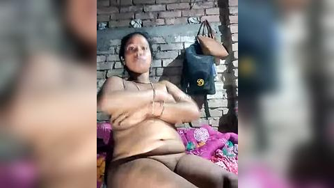 Media: Video of a naked, overweight, dark-skinned woman with long hair, arms crossed, sitting on a bed with pink sheets against a brick wall. She wears only a brown thong. A black handbag hangs nearby.