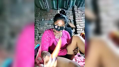 Media: Video of a woman with dark skin, wearing a black mask, pink shirt, and shorts, sitting on a bed in a rustic room with exposed brick walls. She holds a lit cigarette in her outstretched hand, creating a blurred foreground.