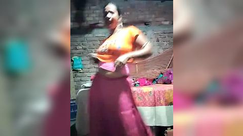 Media: Video of an Indian woman with short hair, wearing a bright orange crop top and pink skirt, dancing in a rustic room with a brick wall, colorful bedding, and a blue object in the background.