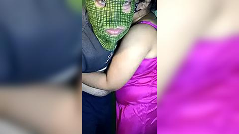 Media: A video depicts a woman in a bright pink dress hugging a man in a green face mask, both standing outdoors. The background is blurred, emphasizing the intimate moment.