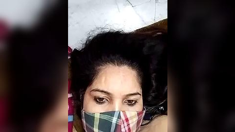 Media: A close-up video of a woman with dark hair, wearing a colorful plaid mask, with a blurred background.