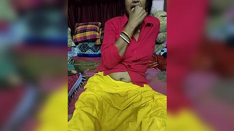 Media: Video of a woman with medium skin tone, wearing a red long-sleeve shirt, yellow sari, and gold bracelets, sitting on a colorful rug with patterned cushions in a dimly lit room.