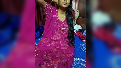 Media: Video of an elderly woman with long black hair, wearing a vibrant pink saree with intricate designs, seated on a blue-patterned bedspread in a dimly lit room.