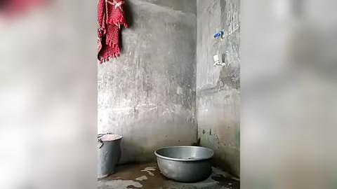 Media: Video of a small, dilapidated bathroom with peeling grey walls, a red towel hanging, a metal bucket, and a large metal basin, indicating a utilitarian, poor living condition.