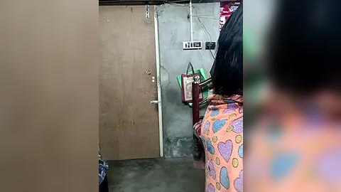 Media: A video of a woman with shoulder-length black hair, wearing a colorful, patterned skirt, standing in a dimly lit, concrete-walled room with a brown door and electrical outlets on the left.