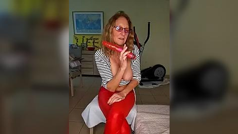 Media: Video of a middle-aged woman with glasses, red lipstick, and red dress, holding a red carrot in a bedroom with a bed, exercise bike, and framed art.