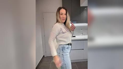 Media: Video of a young woman with long blonde hair, wearing a white sweater and high-waisted light blue jeans, standing in a modern, minimalist kitchen with white cabinets and gray tile flooring.