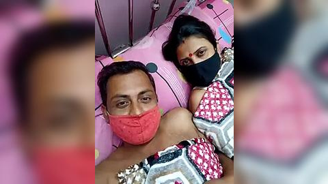 Media: Video of a man and woman lying on a pink, patterned bed, both wearing face masks, with a blurred background, likely in a hospital or medical setting.