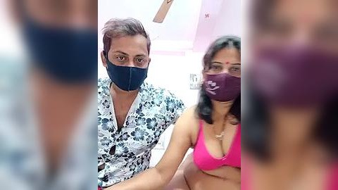 Media: Video of a man and woman wearing face masks, sitting in a pink room; blurred figures in the background.