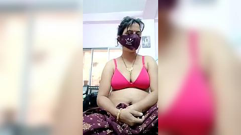 Media: Video of a South Asian woman with medium skin tone, wearing a pink bra, purple mask, and maroon saree, sitting on a bed in a well-lit room with a window.