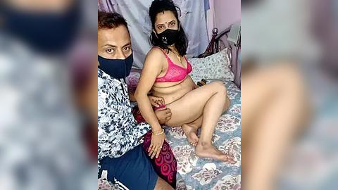 Media: Video of a woman in pink lingerie and black mask being held by a man in a dark room with floral-patterned bedspread.