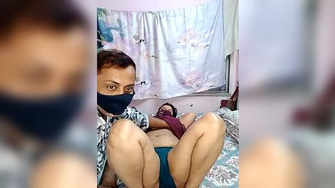 Media: A video of a man in a hospital bed with a blue face mask, looking directly at the camera. His legs are raised, and he wears blue shorts. The room has a white curtain with pastel patterns.