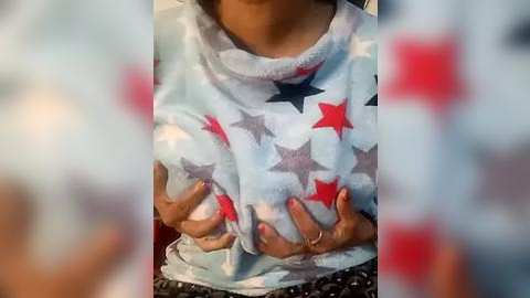 Media: Video of a person wearing a light blue fleece with red and black star pattern, hands on chest, blurred background, indoor setting.