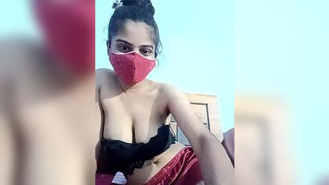 Media: A video of a young woman with light brown skin and dark hair in a bun, wearing a red face mask and black lace bra, seated on a bed with red sheets. The background is blurred, with a blue sky visible.