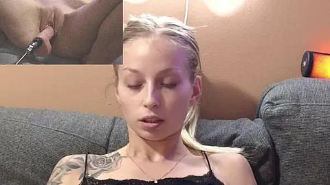 Media: Video of a blonde woman with closed eyes and a tattoo on her shoulder, sitting on a gray couch, holding a vibrator to her vulva.